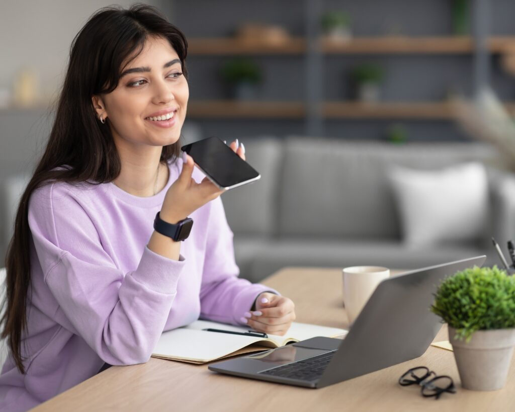 A woman with long dark hair wearing a lavender sweatshirt sits at a desk, smiling while holding her smartphone up as if recording a voice message. A laptop, notebook, small plant, coffee mug, and glasses are arranged on the wooden desk.