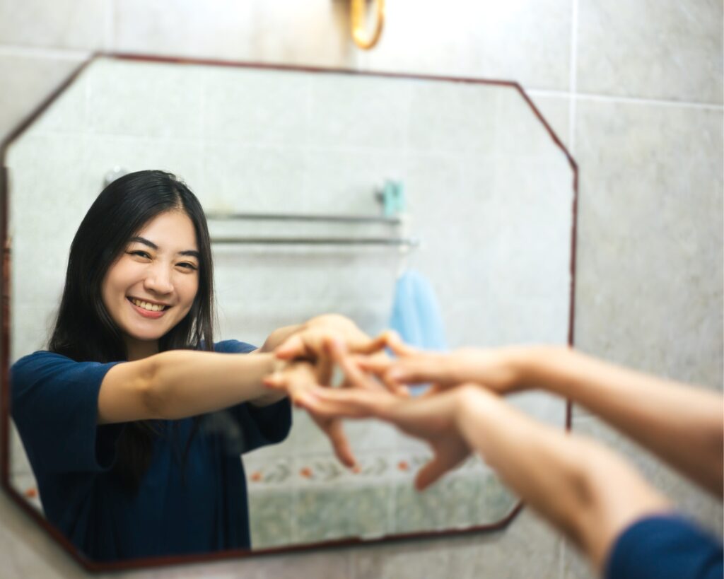 A woman with long dark hair in a navy blue top smiles warmly at her reflection in a bathroom mirror while forming a heart shape with her hands. Both her reflection and her hands reaching toward the mirror are visible.