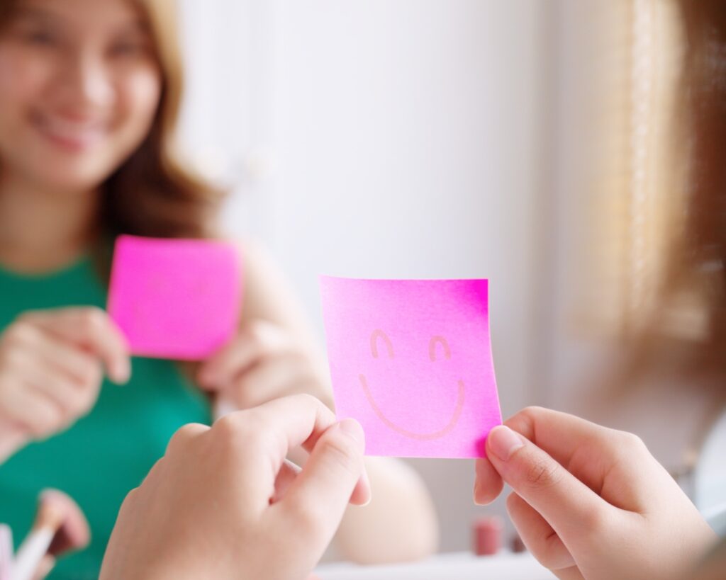 Close-up of hands holding a bright pink sticky note with a simple smiley face drawn on it. In the soft-focus background, another person in a green top holds a similar pink note.