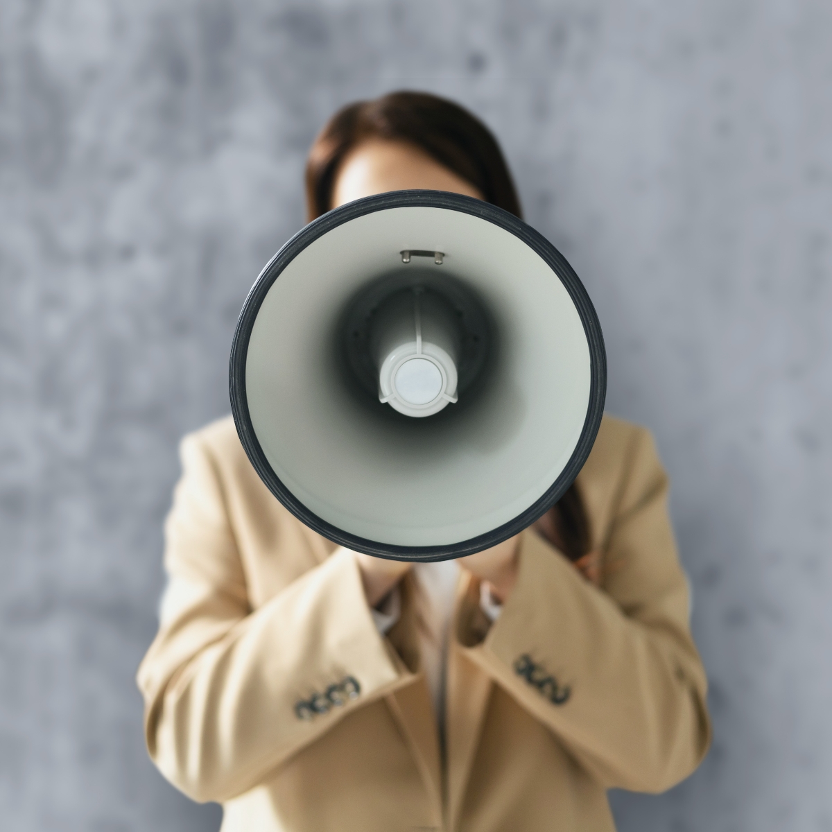 A person in a tan blazer holds a megaphone directly toward the camera, completely obscuring their face. The image shows a straight-on view into the megaphone's cone against a gray concrete wall background.