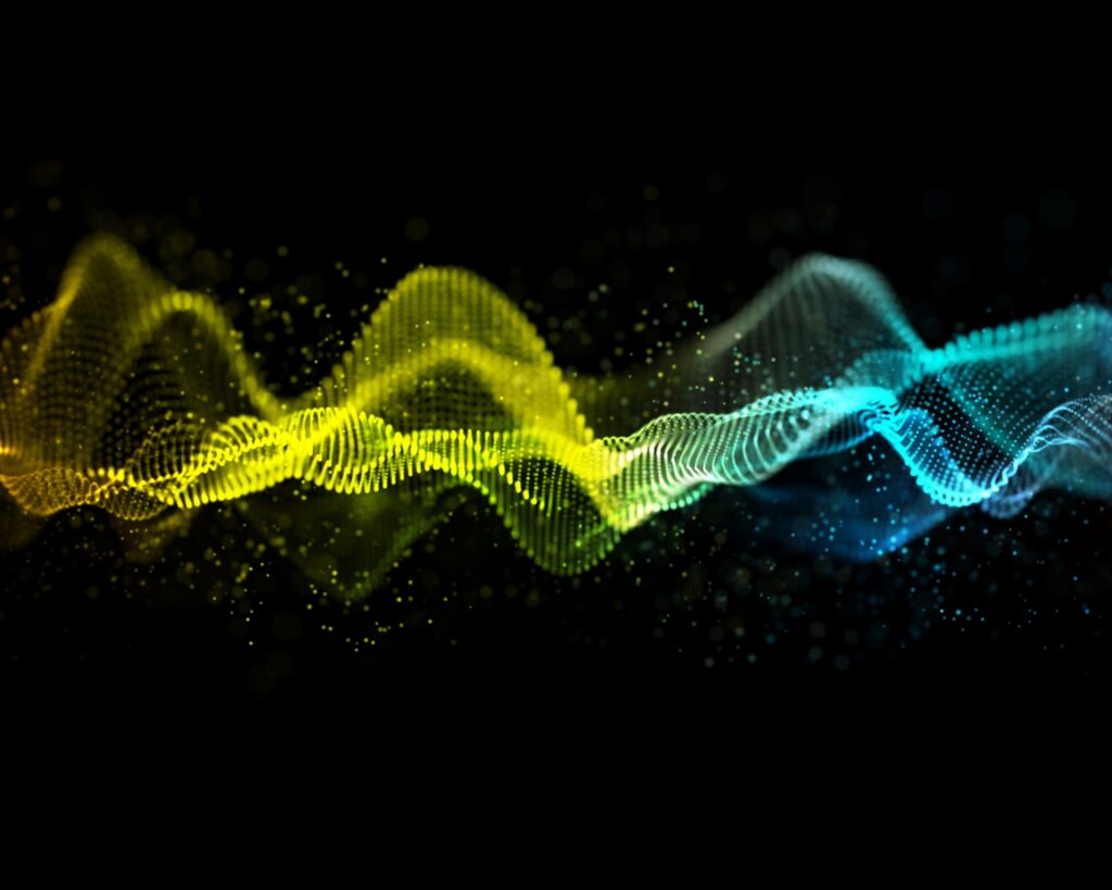 Abstract digital visualization of sound waves made of glowing particles flowing horizontally across a black background. The wave transitions from warm yellow tones on the left through green to cool cyan and blue on the right.