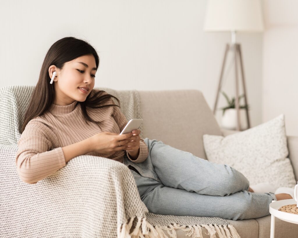 A woman with long dark hair lounges on a gray sofa with a cream-colored throw blanket, wearing wireless earbuds while looking at her smartphone. She's dressed in a beige sweater and light jeans in a minimalist living room with a tripod floor lamp.