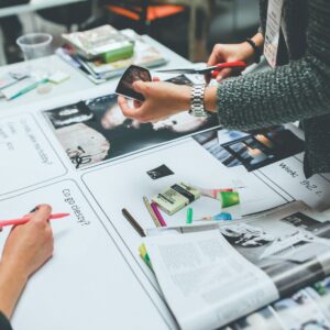 A creative brainstorming session at a white table covered with magazines, photographs, markers, and scissors. One person cuts out an image while another writes on paper worksheets, surrounded by colorful craft supplies and mood board materials.
