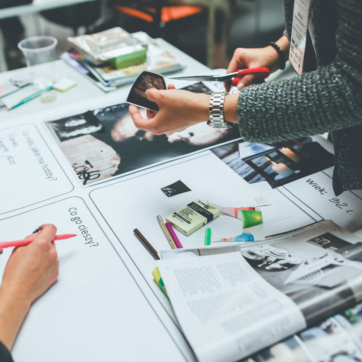 A creative brainstorming session at a white table covered with magazines, photographs, markers, and scissors. One person cuts out an image while another writes on paper worksheets, surrounded by colorful craft supplies and mood board materials.