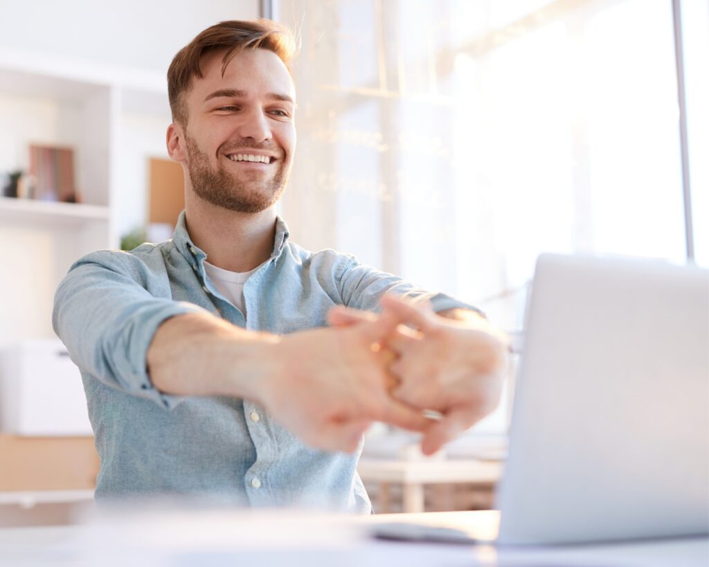 Man smiling and stretching at his desk in front of a laptop, preparing for a productive and creative workday.