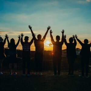 Silhouettes of seven people standing in a row with their arms raised triumphantly against a golden sunset sky. The sun is low on the horizon, creating a warm glow and lens flare behind the central figure.