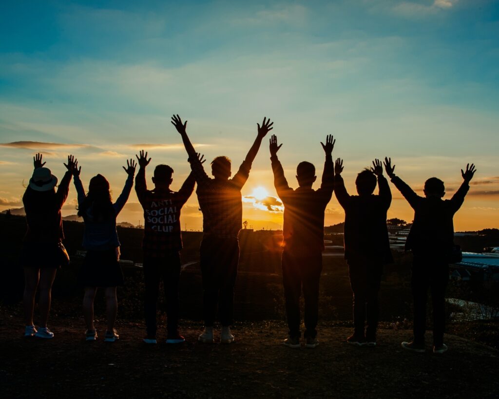 Silhouettes of seven people standing in a row with their arms raised triumphantly against a golden sunset sky. The sun is low on the horizon, creating a warm glow and lens flare behind the central figure.