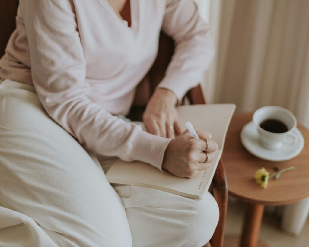 A woman in a soft pink sweater and cream-colored pants sits writing in an open journal resting on her lap. A wooden side table holds a white cup of black coffee on a saucer, with a small yellow flower beside it. The image is cropped to show her torso and hands, with warm, muted tones throughout.